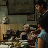 A group of people is gathered around a table, sharing a meal. The table is set with various dishes, including a plate of fish, a bowl of soup, and several smaller bowls containing different foods. The setting appears to be a modest home with a mix of traditional and modern elements, including patterned wallpaper, framed pictures, and a large painting on the wall. A shelf with books and containers is also visible in the background.