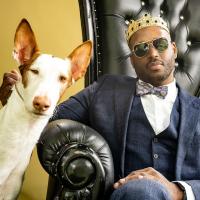 Shane Campbell-Staton with crown on his head sitting in a chair shaped like a throne leaning towards a tall skinny dog that's leaning towards him. 