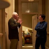 An older man talking to a woman who stands next to him with her arms crossed. There is a lamp in the foreground of the shot.