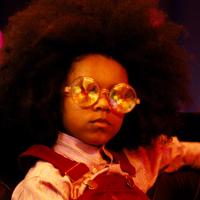 A mysterious looking young girl with an afro wearing reflective round sunglasses and overalls.