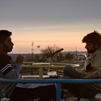 Two men sit facing each other with a sunset and construction debris in the background.