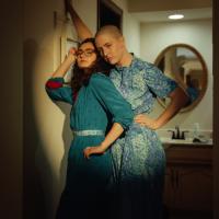 Two women -- one with a shaved head and one with long curly hair and glasses -- pose in front of a bathroom.