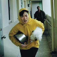 A person wearing a yellow rain jacket is being chased down a hallway carrying a large cooking pot and a small white dog.