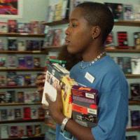 A grainy image of woman with a blue shirt carrying an armful of VHS tapes inside a video store.