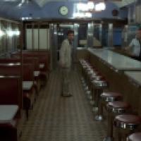 Interior of a classic American diner with red leather booths and red cushioned stools at a counter; a man in a light suit stands facing a uniformed man behind the counter, with a clock and menu boards visible in the background