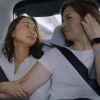 Two women in white shirts embrace in the back of a car wearing seatbelts.