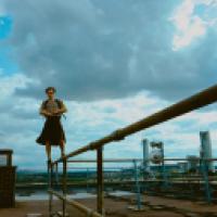 Person standing on a metal railing on a rooftop, with an industrial area and cloudy sky in the background