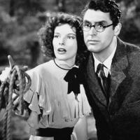Two people stand close together outdoors; one wears a ruffled dress and holds a coiled rope, the other wears a suit and tie