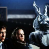 Three individuals sit in a dimly lit setting; one wears a large rabbit costume with sharp teeth and prominent ears