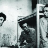 Three individuals in a prison room with bunk beds and graffiti on the wall; one wears a uniform labeled '19421'. Two cups sit on a nearby surface