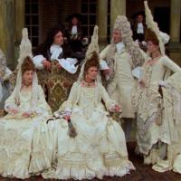 Group of people in ornate 17th or 18th-century costumes with wigs, posed in front of a columned building