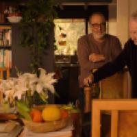 Two people stand near a table holding a basket of fruits, white lilies in a vase, and papers. A bookshelf filled with books and plants is visible in the background