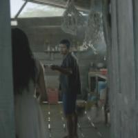 Inside a dimly lit rustic room with wooden walls and floor, one person stands holding an object while another is partially visible in the foreground; ropes hang from the ceiling and shelves line the back wall