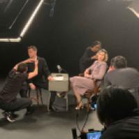 Behind-the-scenes setup of a photoshoot or video recording with two seated individuals and a microphone between them. Crew members adjust lighting and equipment in a dark setting