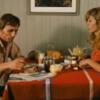 Two people sit at a breakfast table with a red, white, and blue striped tablecloth, surrounded by cups, a teapot, jam, juice, bread, and a newspaper; striped wallpaper and a framed picture are in the background