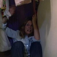 A frightened person sits hiding on the floor of a closet surrounded by hanging clothes