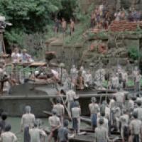 A movie set scene with a boat carrying crew and equipment in the water, surrounded by people in small boats or standing in water, many covered in white paint; an ancient stone structure with vegetation is in the background