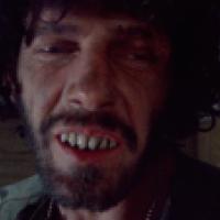 Close up image of a person's face with black curly hair, facial hair, and exposed teeth.