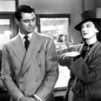 Black and white photo of two individuals in an office setting; one wears a double-breasted suit with a tie, the other a striped outfit with a hat