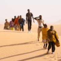 Ten individuals walk in a line across a desert landscape with rolling sand dunes under a clear sky