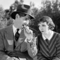 Two people sit outdoors among trees; one wears a suit, tie, and hat while biting a carrot, the other wears a striped dress with a white collar