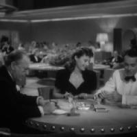 Three people sit at a round table in a casino or upscale club, with drinks and an ashtray; other patrons and ambient lighting are visible in the background