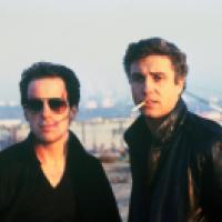 Two individuals stand outdoors against an industrial or urban backdrop; one wears a dark shirt and the other a dark jacket