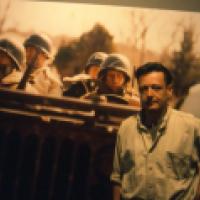 A large photo image of a group of people, some in helmets and uniforms, stand behind a barricade while a casually dressed individual stands in the foreground