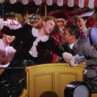 A group of people in late 19th or early 20th century costumes ride on an open carriage with a yellow front panel and decorative ironwork