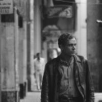 The image is black and white. A man in a leather jacket walks down an urban street with buildings, signs, and other pedestrians in the background.