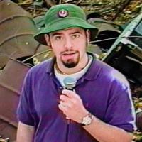 A person in a green hat and purple shirt holds a microphone in front of rusted metal barrels and debris.