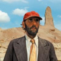 A person in a red cap, suit jacket, and orange tie stands in a desert landscape with rock formations in the background.