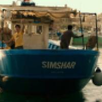 A blue fishing boat named 'SIMSHAR' is docked at a busy harbor with three people aboard; other boats and buildings are visible in the background