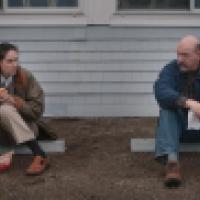 Two people sit on the ground outside a building; one is eating a sandwich while the other holds a napkin
