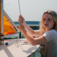 A young woman with long blonde hair sails a small sailboat with an orange sail on a lake.