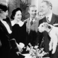 Black and white photo of four people in formal attire; the man on the right holds a small dog
