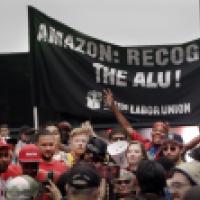 Group of people holding a large black banner that reads 'AMAZON: RECOGNIZE THE ALU!' with 'AMAZON LABOR UNION' and raised fists printed below