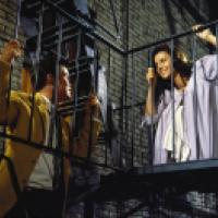 Two people stand on different levels of a fire escape attached to a brick building. One wears a brown jacket on the lower level, while the other in a light purple dress looks down from the upper level