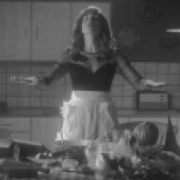Person with arms outstretched stands in a kitchen wearing a black top with sheer sleeves and a white apron. The kitchen features tiled walls, cabinets, appliances, and a table with food items like bread, fruit, and baked goods