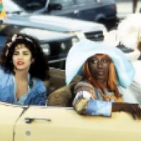 Three individuals sit in a yellow convertible; one wears a blue shirt, another a large blue hat and colorful clothing, and the third a white outfit with a white hat