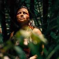 Person standing in a dense forest with lush green foliage; their face is partially covered by leaves and branches
