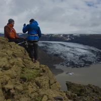 Two people in ski jackets perched on a mountain cliff high above melting glaciers and a lake.