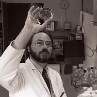 A black and white still image of a man in a white lab coat holding a petri dish up above his face to examine it.