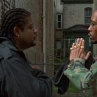 A man wearing a black jacket stands facing a man with his hands in a prayer gesture wearing a camo jacket and lots of gold rings.
