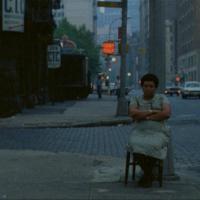A woman sitting with her arms crossed in a wooden chair at a city intersection. There is a "Don't Walk" sign illuminated above her.