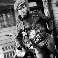 A man with a leather jacket and military cap leaning casually against a motorcycle.