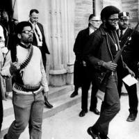A group of young men exiting a building carrying rifles. The man at center is wearing bullets across his chest and around his waist.