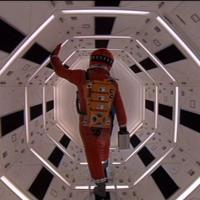 A person in a red space suit walks through a brightly lit, hexagonal corridor with futuristic design.