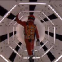 A person in a red space suit walks through a brightly lit, hexagonal corridor with futuristic design.
