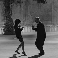 Two people dance energetically in an open courtyard, one in a dress and heels, the other in a suit. A third person sits at a table under a canopy, watching the scene unfold.